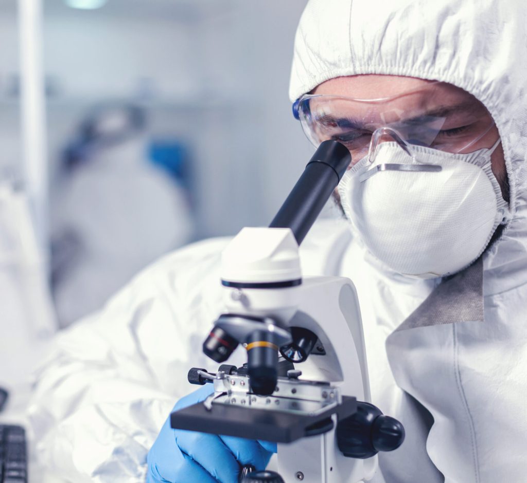 Chemical engineer wearing glasses conducting health investigation on microscope. Scientist in protective suit sitting at workplace using modern medical technology during global epidemic.