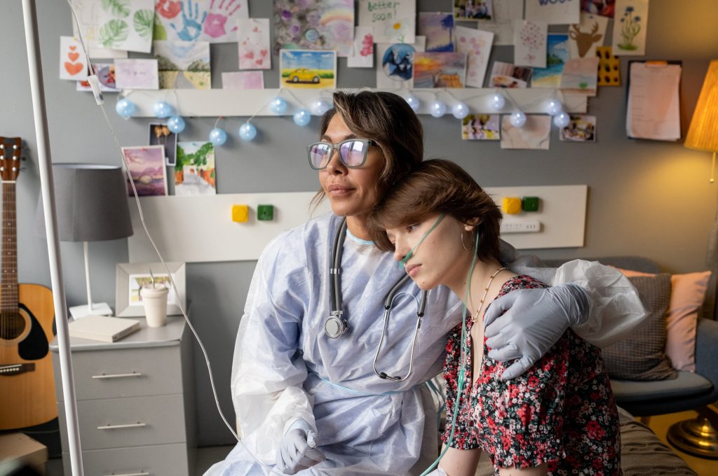 Nurse in workwear and eyeglasses embracing teenage patient with oxygen nose pipe while sitting next to her in ward
